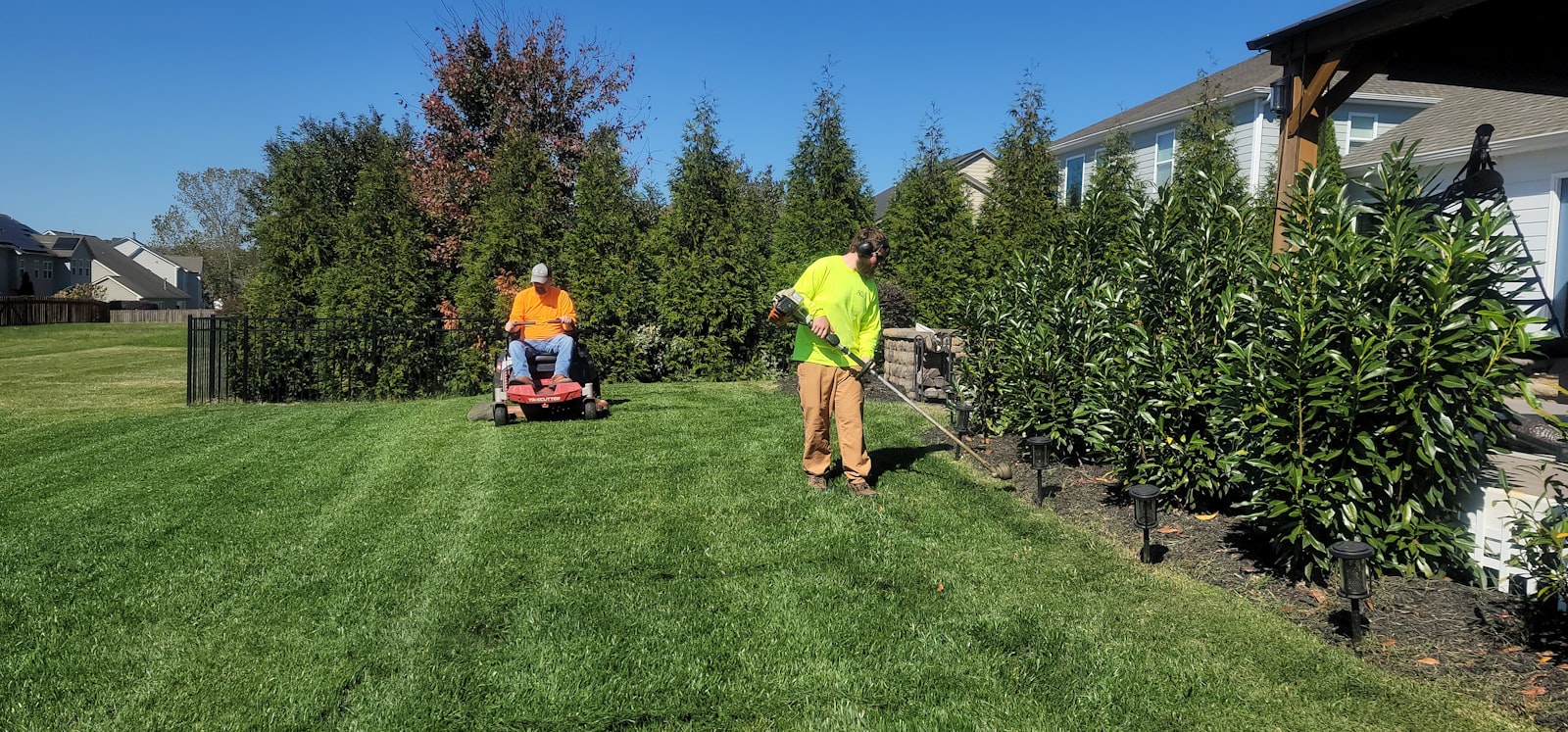 Solo operator pushing a walk-behind mower along the side yard of a brick home.